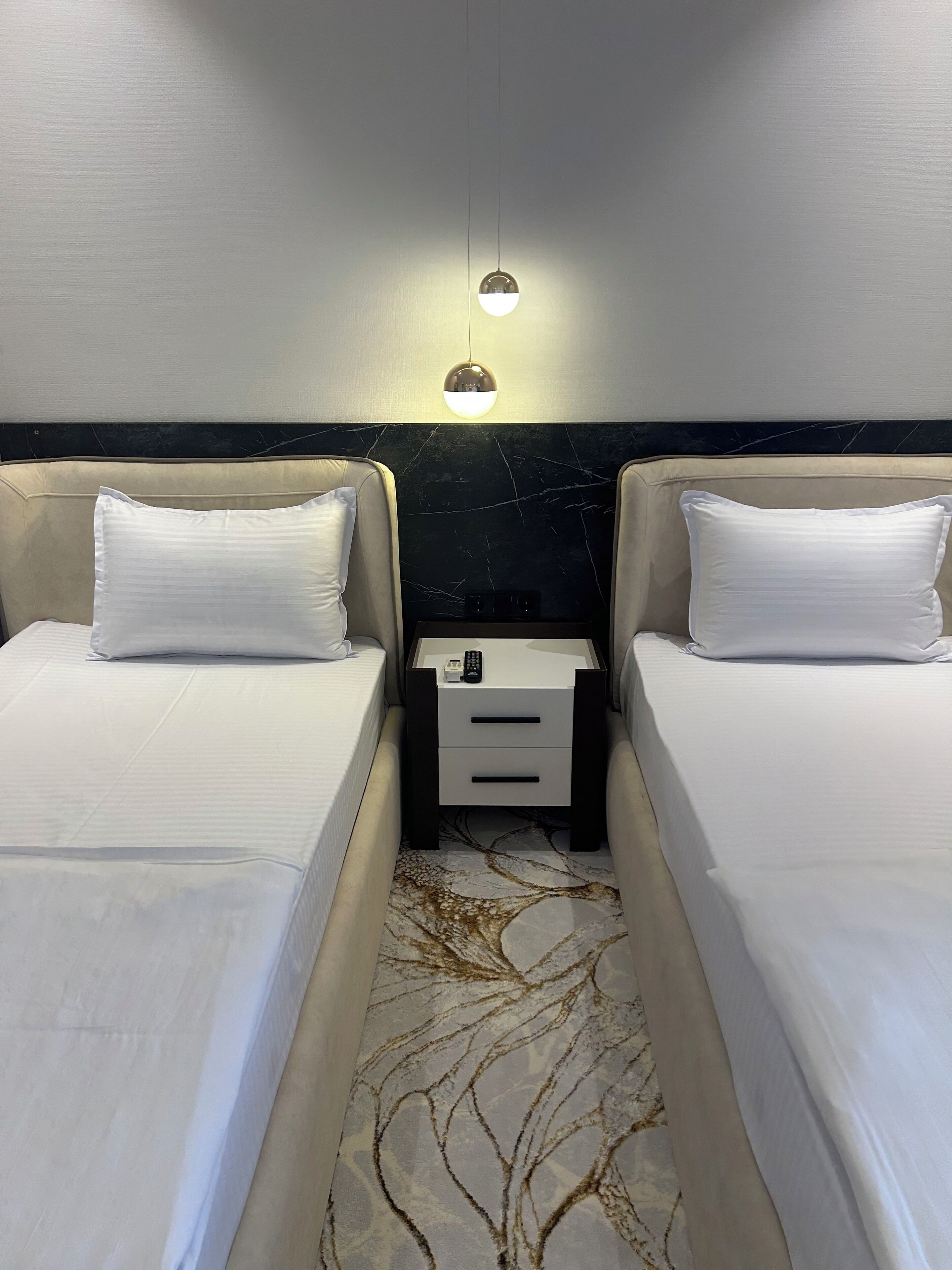 A hotel room with two single beds, a nightstand with a lamp, and a marble-patterned floor.