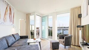 A modern living room with a grey sofa, a small table, a grey armchair, and a TV on a stand. The room has large windows with a view of a marina and a bridge. - HighEnd Travemünde - Whg 404 (Lübeck Travemünde)