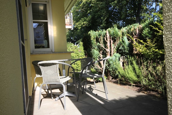 En terrasse med to metalstole og et bord, et vindue med udsigt til træer, og et lille haveområde.