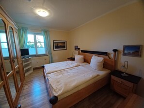 Un lit double avec une tête de lit en bois, deux tables de chevet avec des lampes, une fenêtre avec des rideaux, un bureau avec un ordinateur et un radiateur.