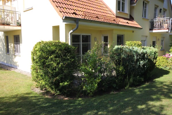 Un bâtiment jaune avec un toit recouvert de tuiles rouges, de nombreuses fenêtres et un petit jardin agrémenté d’arbustes.