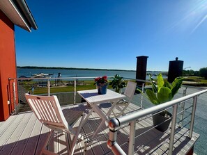 Un balcon avec une table et des chaises, donnant sur une étendue d’eau où l’on peut voir des bateaux et un quai.