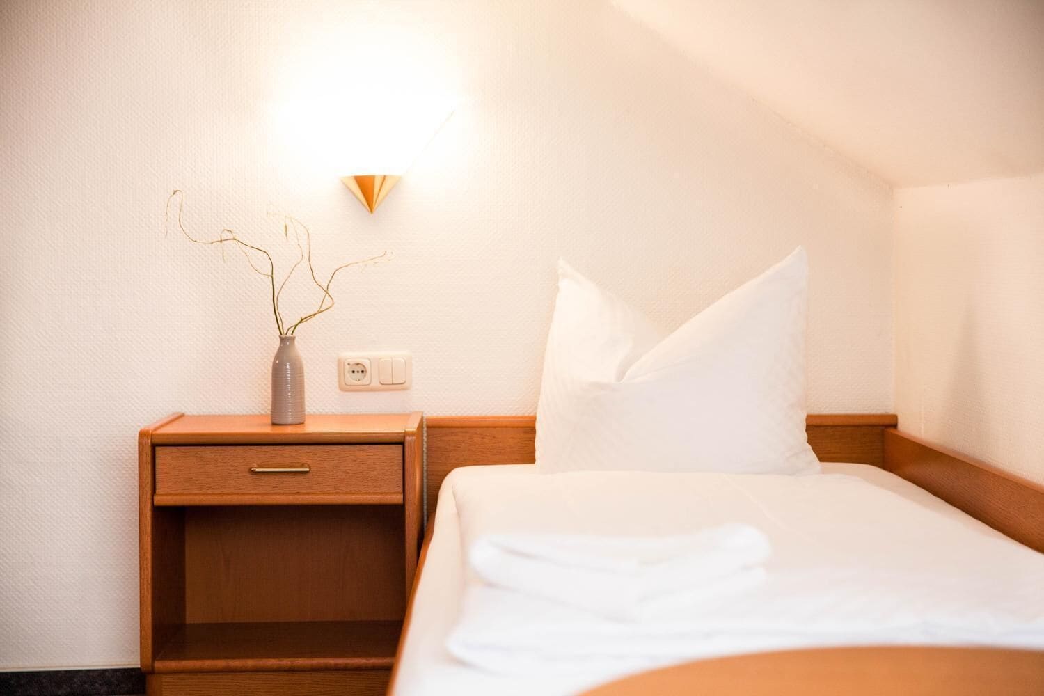 A hotel room with a wooden nightstand, a vase with branches, a bed with white bedding, and a wall-mounted light.