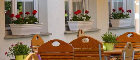 A patio with wooden chairs and tables, potted plants, and a white wall with windows.