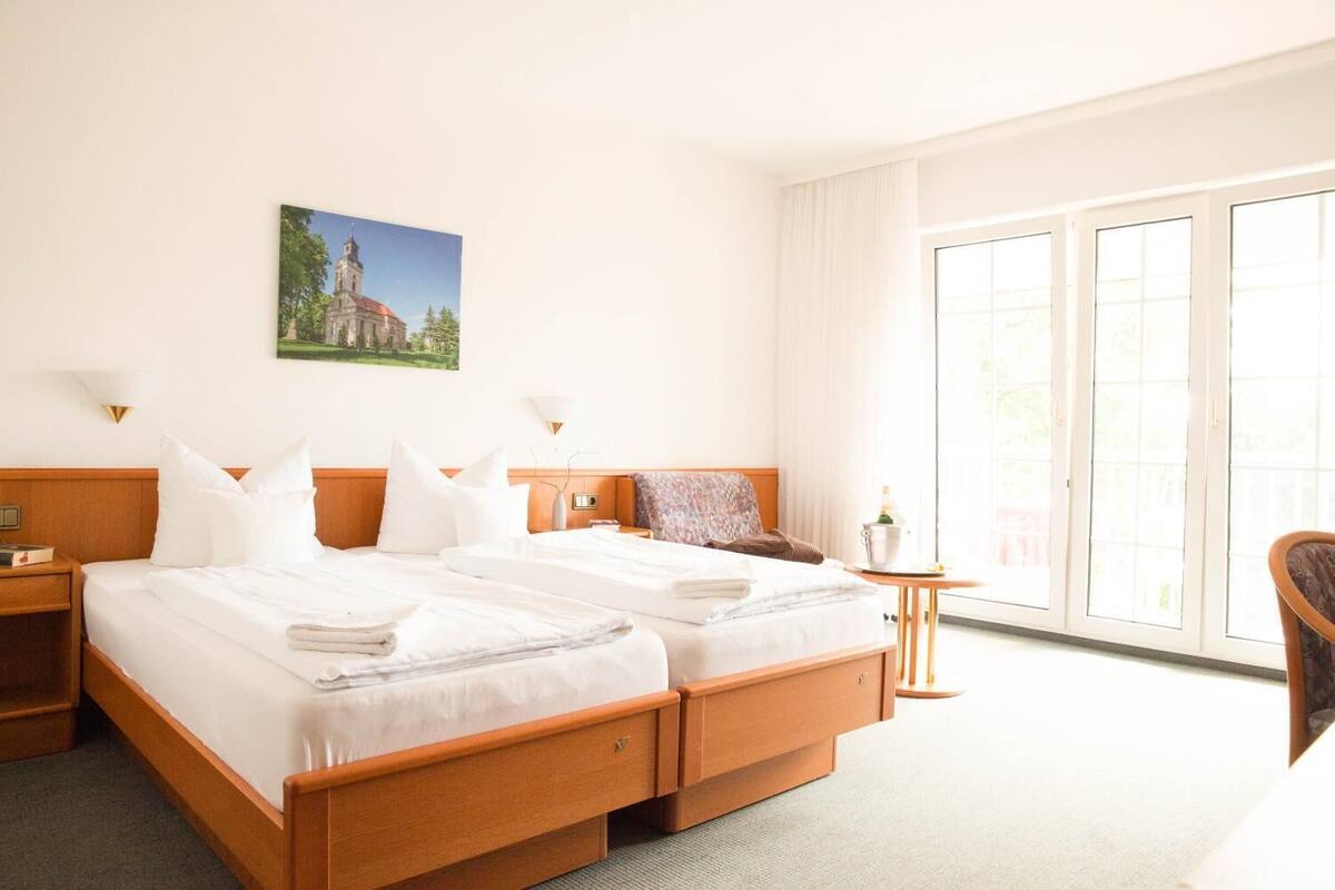 A hotel room with two beds, a nightstand, a chair, a small table, and a large window with a view of a building.