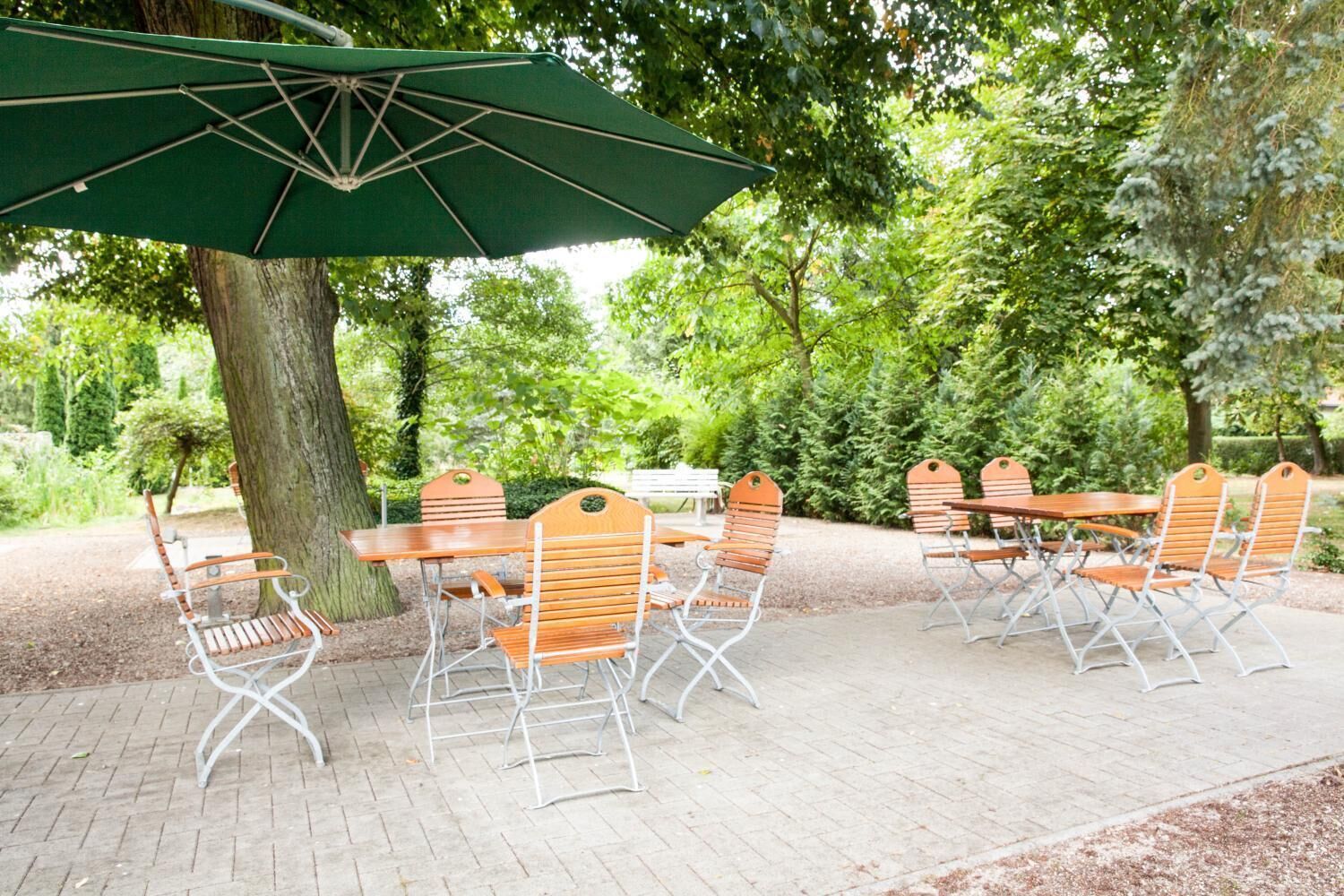 An outdoor patio with a brick floor, wooden tables and chairs, and a green umbrella.