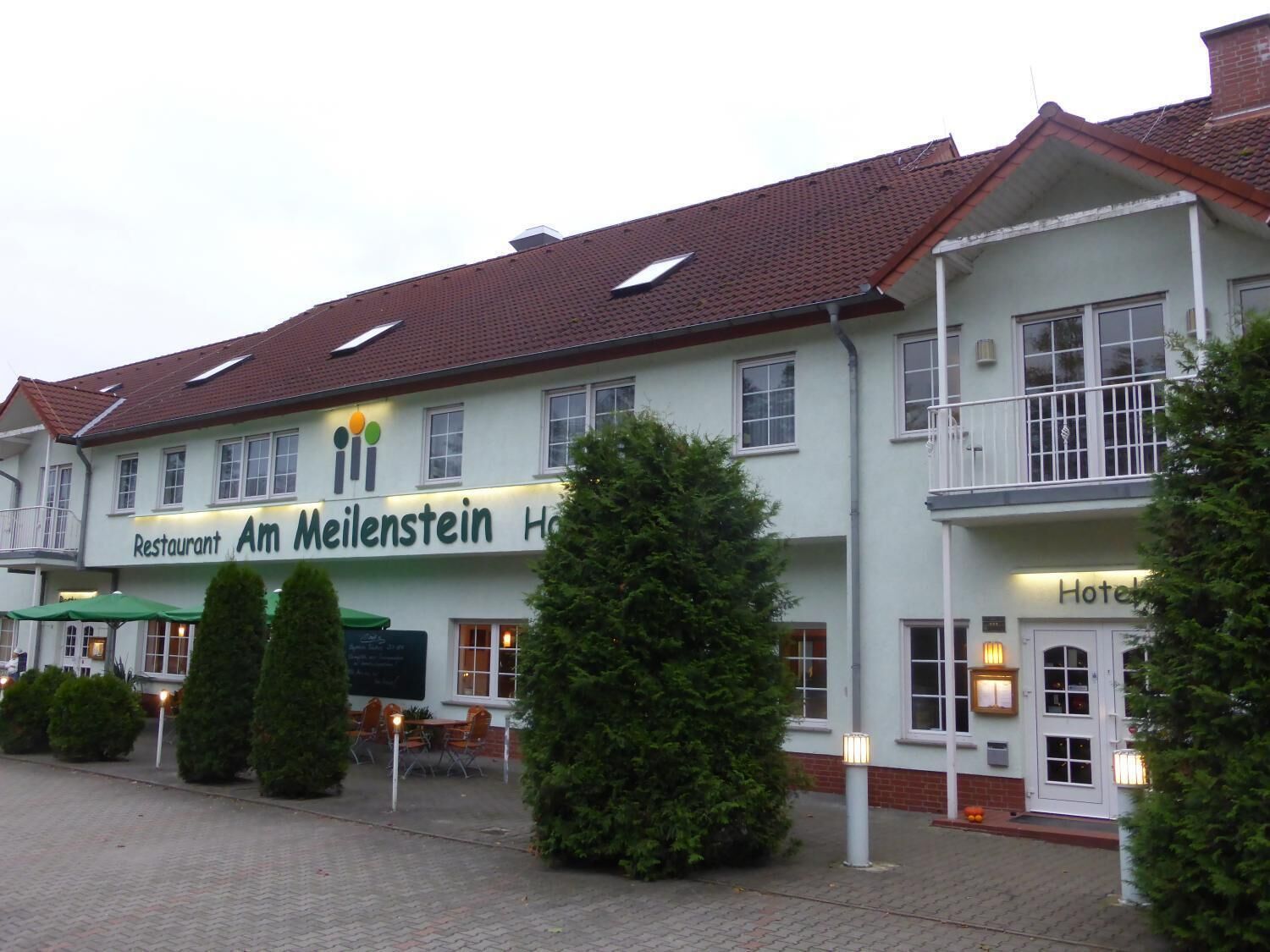 A two-story building with a sign for 'Restaurant Am Meilenstein Hotel'. There are outdoor seating areas with tables and chairs, and the building has a red-tiled roof.