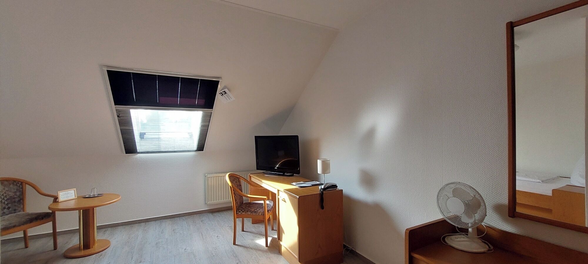 A small, well-lit room with a desk, chair, television, and a mirror.