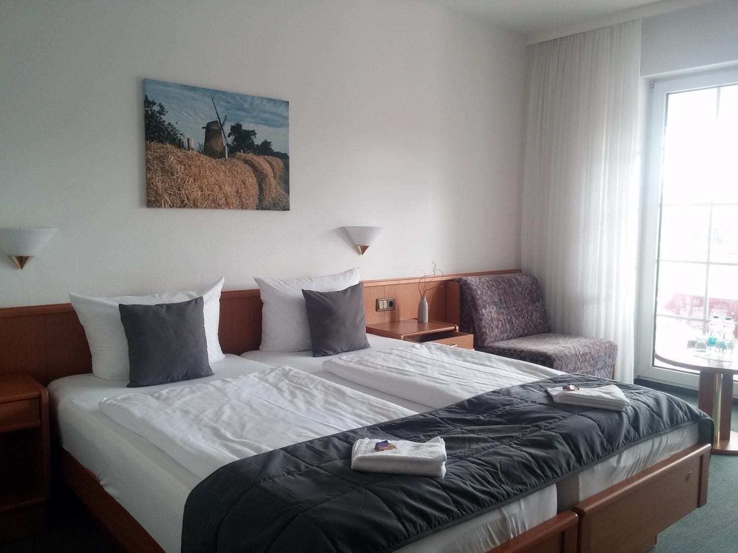 A hotel room with two beds, a large window, a painting on the wall, and a small table with a lamp.