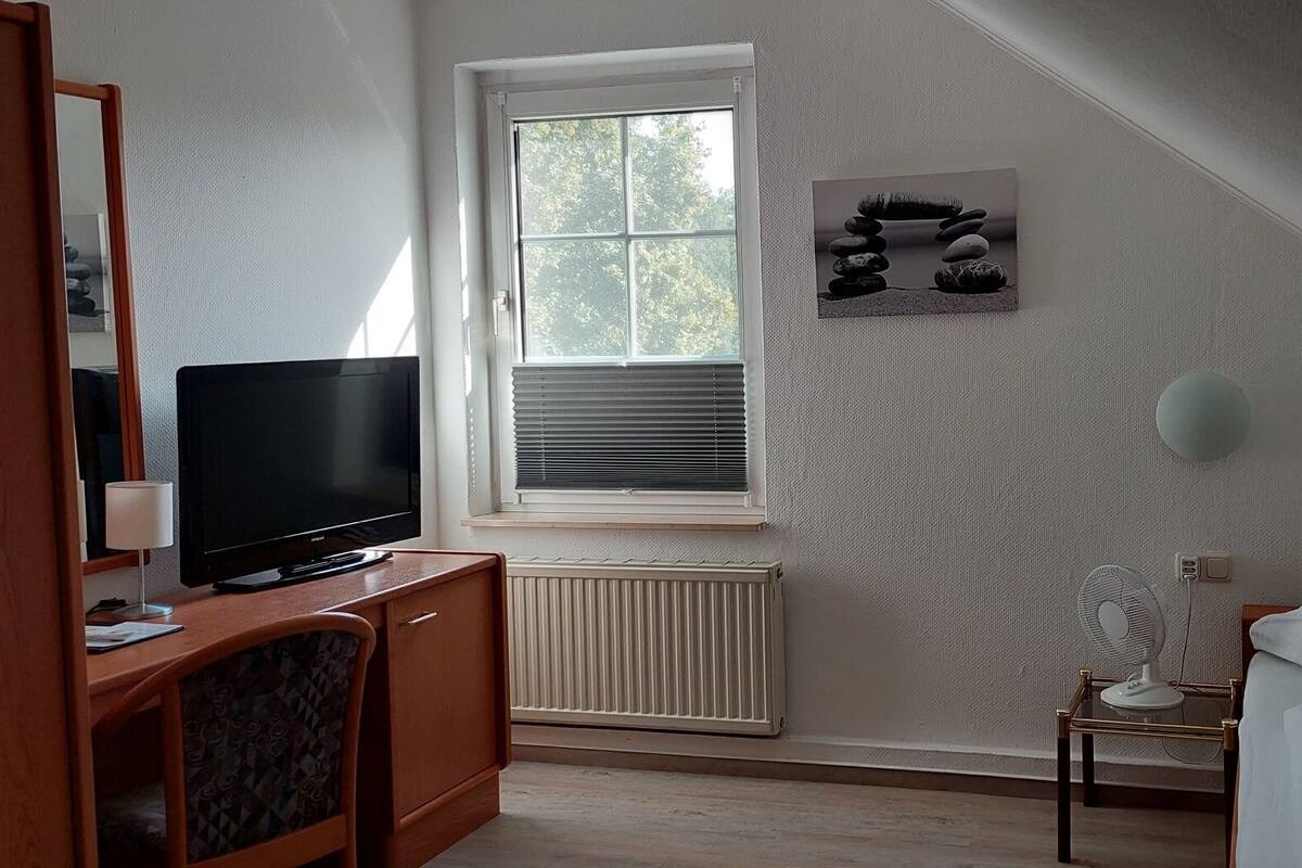 A bedroom with a TV, a bed, a radiator, a window, and a wall-mounted fan.
