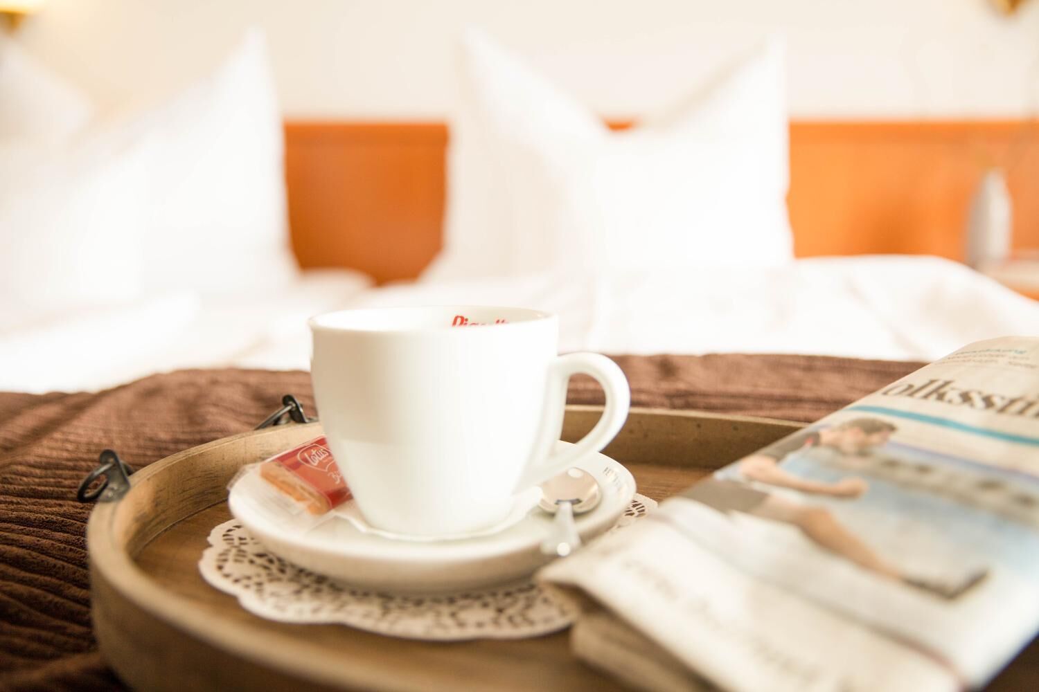 A tray with a white cup and saucer, a tea packet, and a newspaper on a bed with white pillows and an orange headboard.
