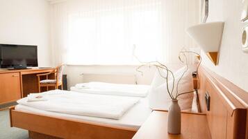 A hotel room with a bed, a desk with a television, a chair, and a bedside table with a vase.