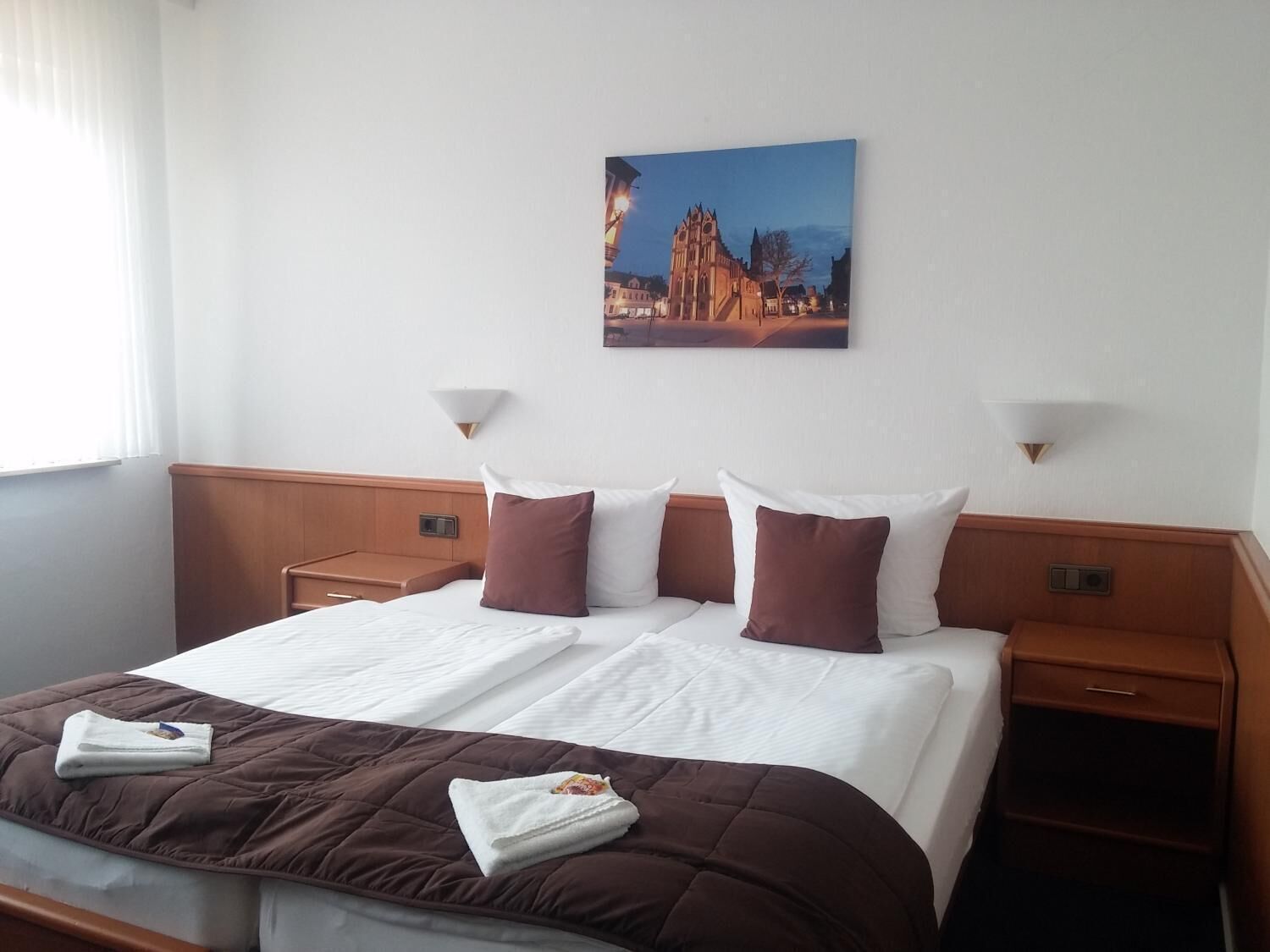 Ein Hotelzimmer mit einem großen Bett, zwei Nachttischen, einem Wandbild und einem Fenster mit Jalousien.