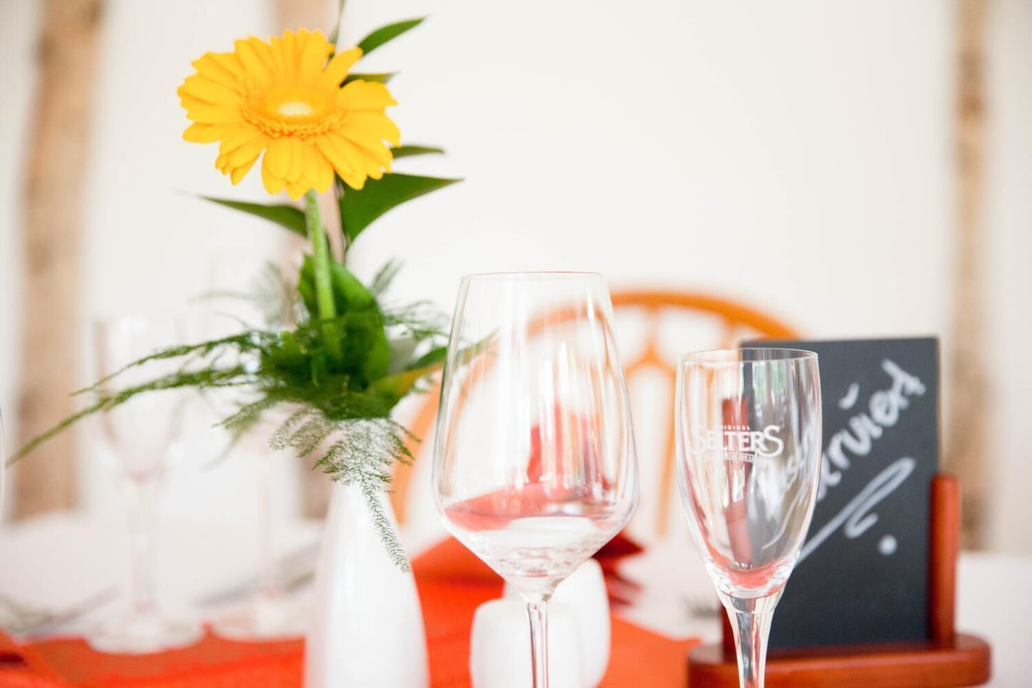 A table with two wine glasses, a yellow flower, and a blackboard with writing.