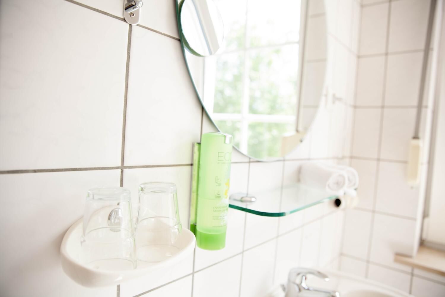 Un baño con azulejos blancos, un estante de vidrio con una botella verde y un espejo.