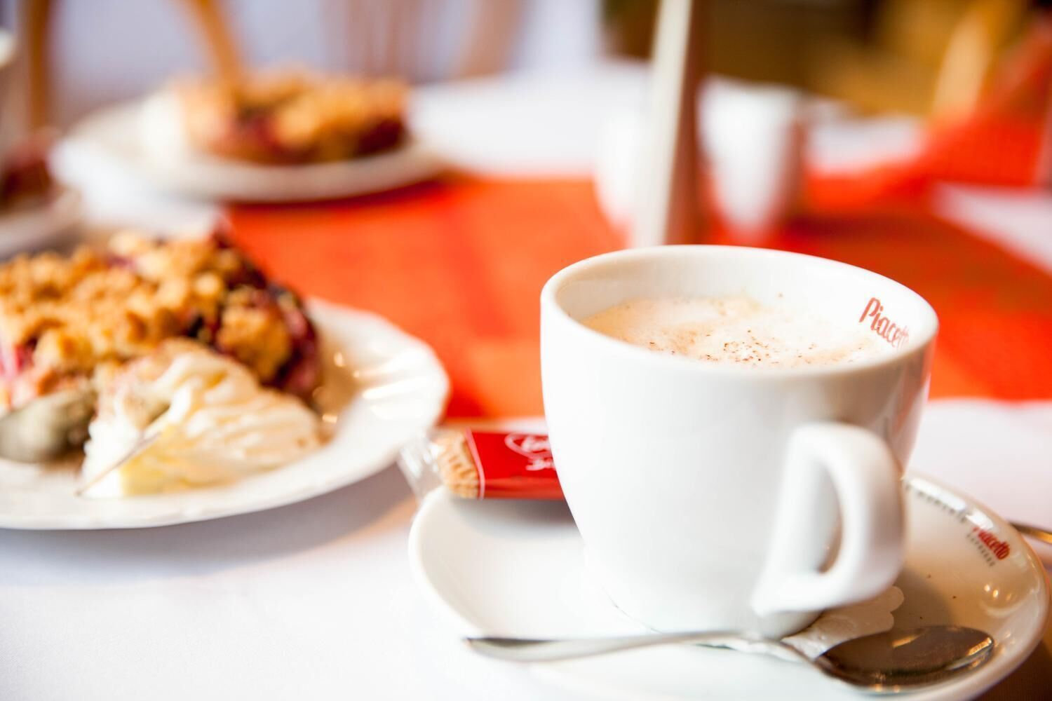 A white cup with a spoon on a saucer, a plate with food, and a red napkin on a table.