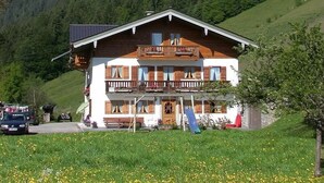 Un chalet de dos plantas con balcón, rodeado de un paisaje verde exuberante y montañas al fondo.
