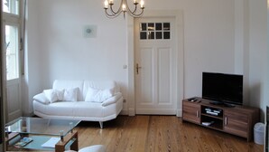 A living room with a white sofa, a glass coffee table, a wooden cabinet with a TV, and a chandelier.