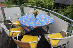 Una mesa redonda con mantel floral, rodeada de sillas de mimbre con cojines amarillos, en un balcón con barandilla.
