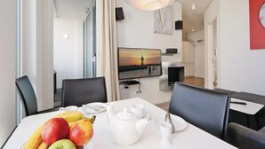 Un comedor moderno con una mesa blanca dispuesta para dos personas, que incluye un frutero, tetera, tazas y un cuadro con un atardecer sobre un muelle.