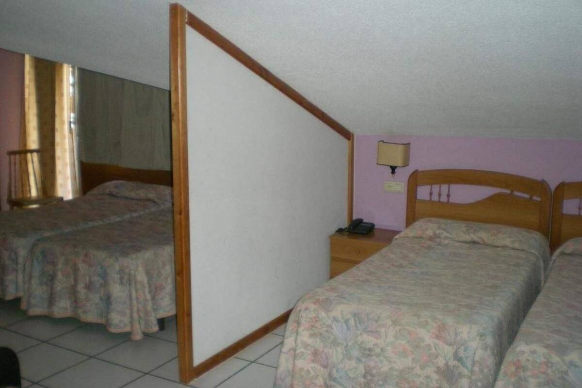 Habitación con dos camas individuales, un separador de madera, una mesita de noche con lámpara y un ventanal con cortinas.