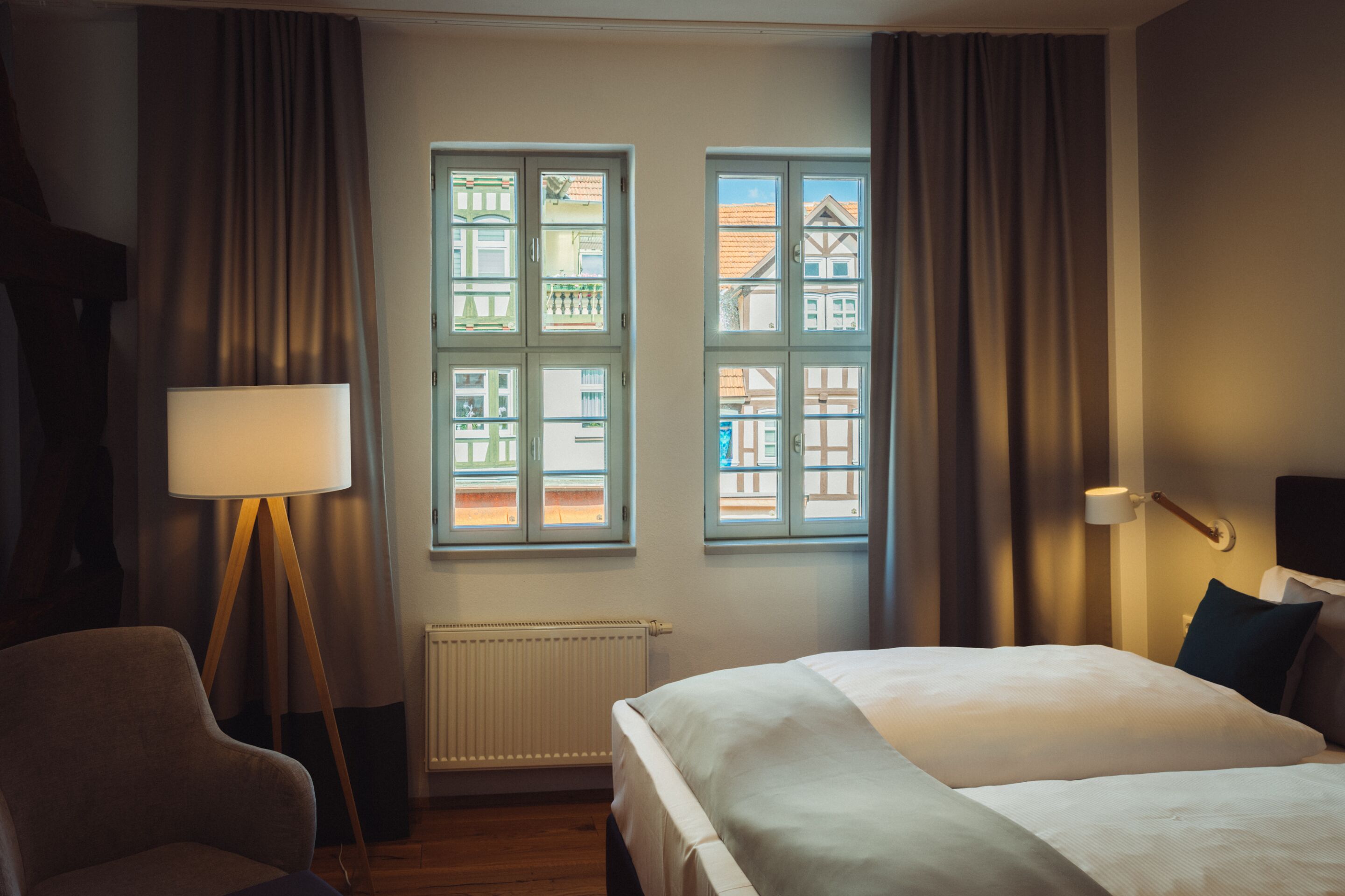 Een hotelkamer met een bed, een stoel, een lamp en twee ramen met uitzicht op gebouwen.