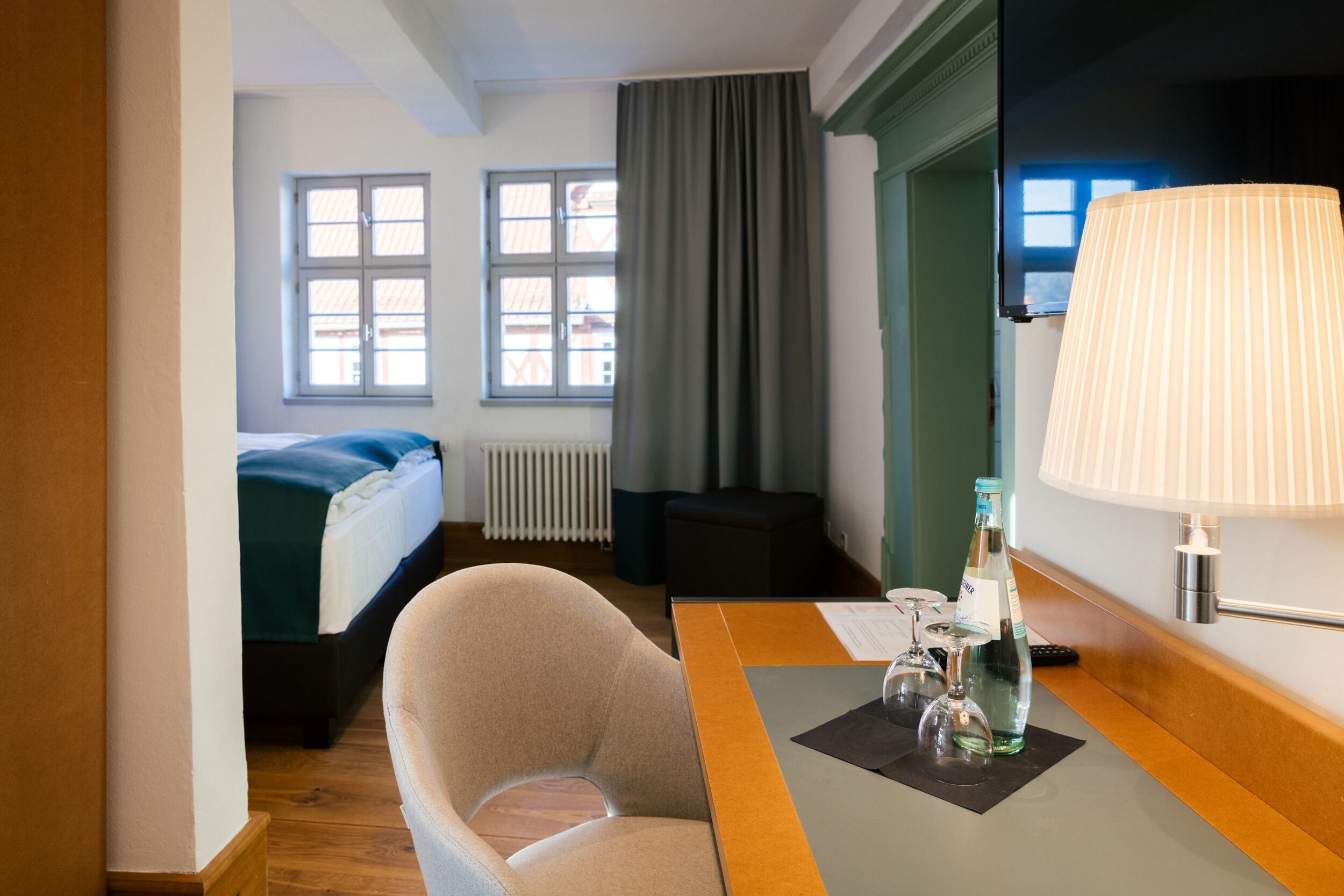 Une chambre d’hôtel avec un lit, une chaise, une table avec une lampe et une fenêtre avec des rideaux.