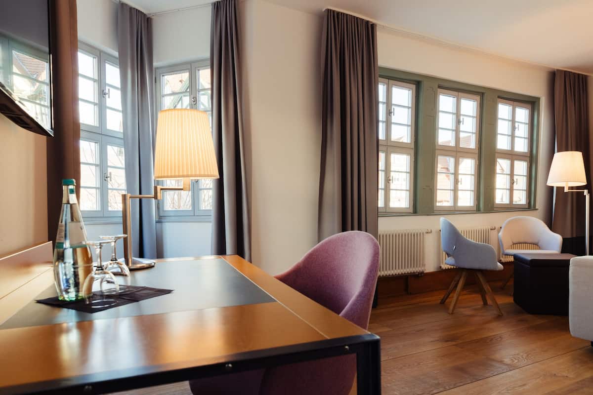 Ett hotellrum med ett träbord, en lila stol, en vit stol, en lampa och ett fönster med gardiner.