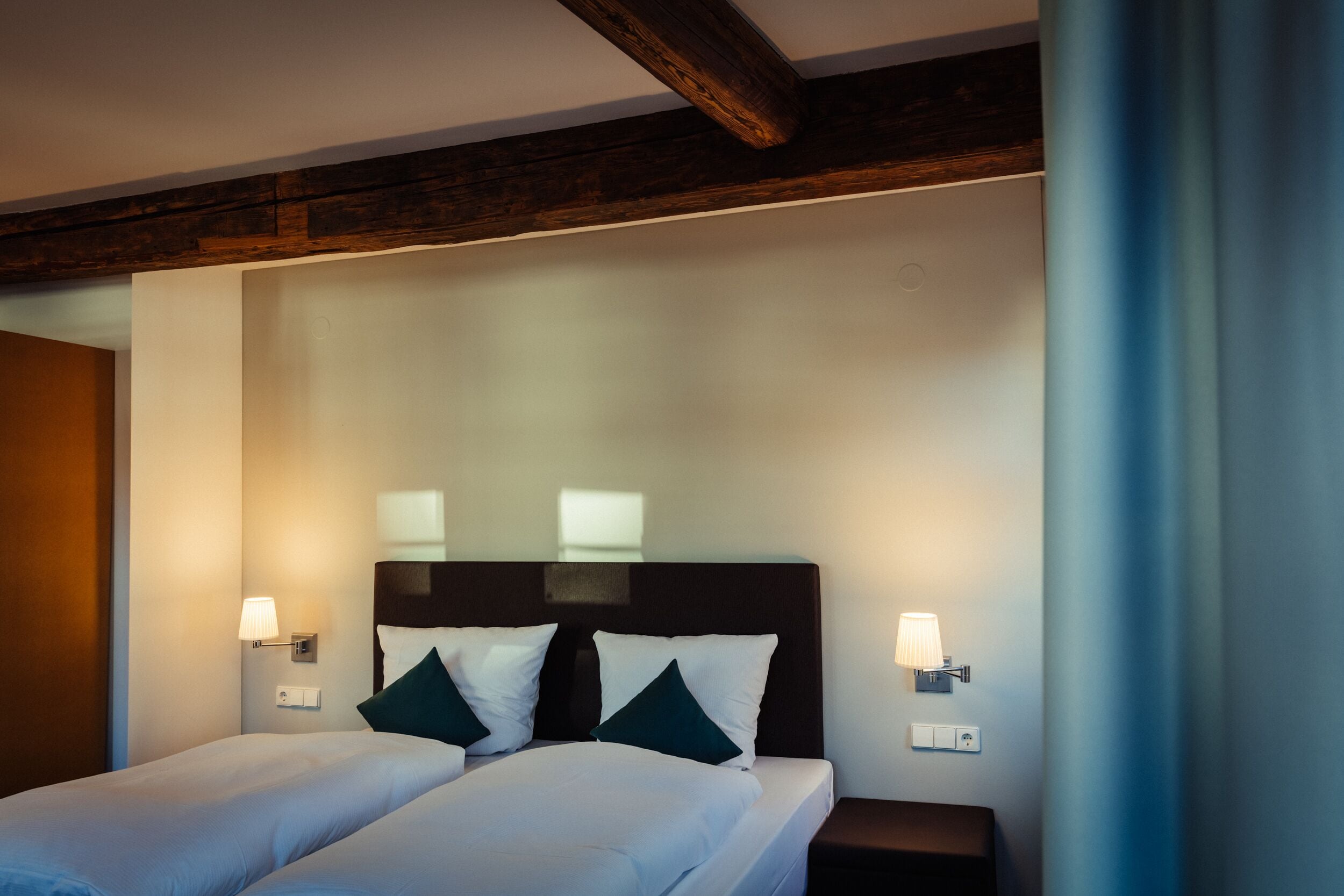Ein Hotelzimmer mit zwei Betten, dunklen Kopfteilen, Nachttischlampen und einem Holzbalken an der Decke.