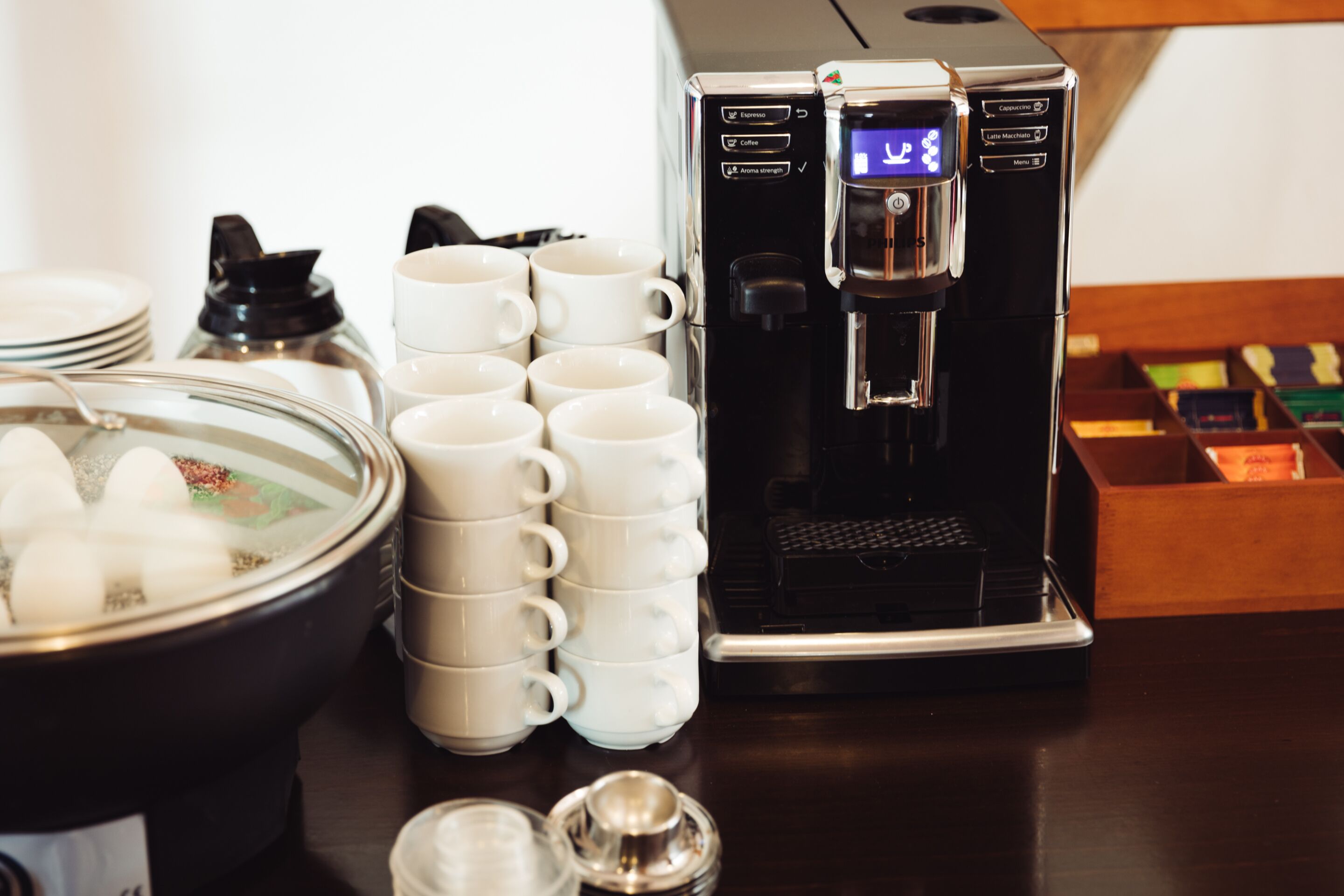 A coffee machine with a digital display, a stack of white cups, and a bowl of ice cubes on a wooden table.