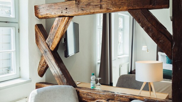 A modern room with exposed wooden beams, a grey armchair, a wooden table, and a TV mounted on the wall. - Hotel Zum Schwan (Wanfried)