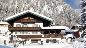 Un bâtiment de style chalet, avec une façade en bois et des volets verts, entouré d’arbres couverts de neige et sous un ciel dégagé.