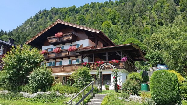 Un chalet traditionnel doté de nombreux balcons ornés de fleurs, entouré d’une végétation luxuriante et surplombant un paysage montagneux.