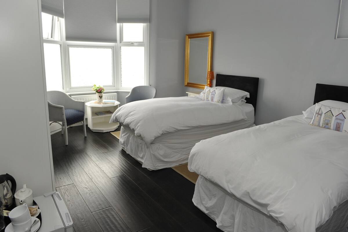 A hotel room with two beds, a small table with a lamp, a chair, and a window with blinds.