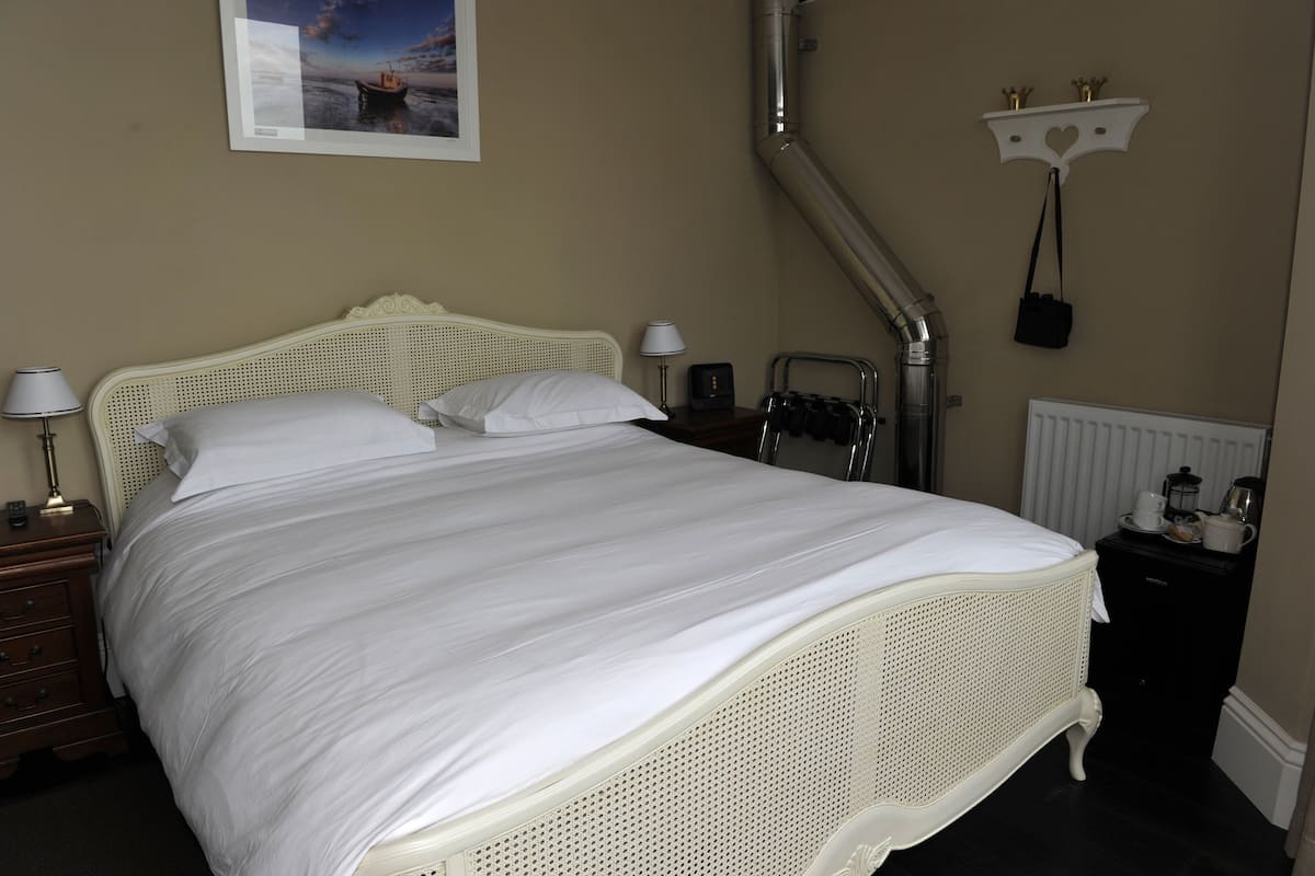 A neatly made bed with white linens, a bedside lamp, a nightstand, a radiator, and a shelf with a small item.
