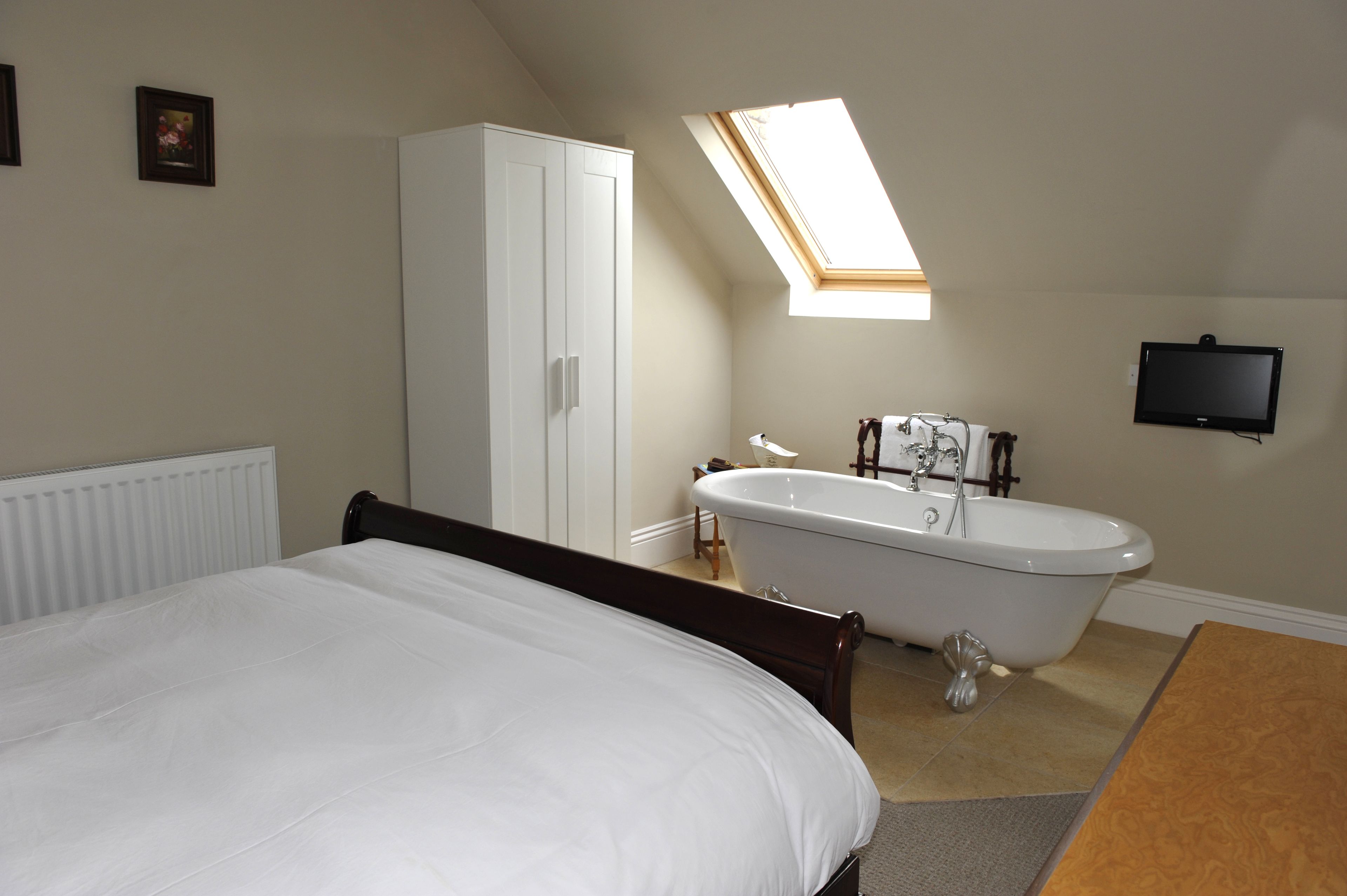 A bedroom with a bed, a white wardrobe, a clawfoot bathtub, a skylight, and a TV mounted on the wall.