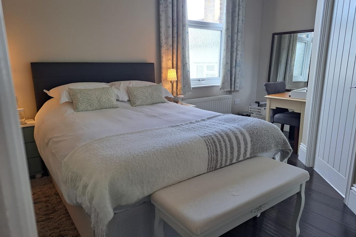 A neatly made bed with a headboard, a bedside table with a lamp, a bench, and a window with curtains.