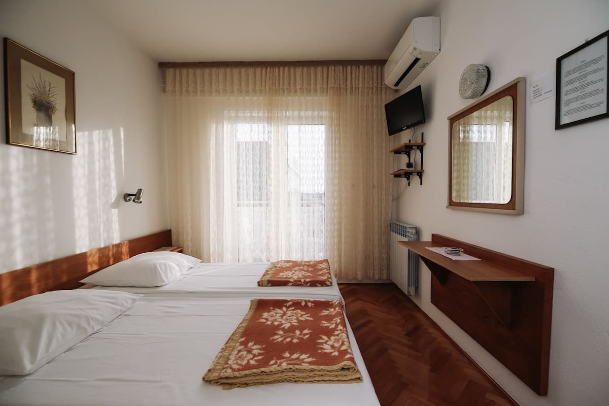 Habitación de hotel con dos camas, cabecera de madera, escritorio, televisor y ventana con cortinas translúcidas.
