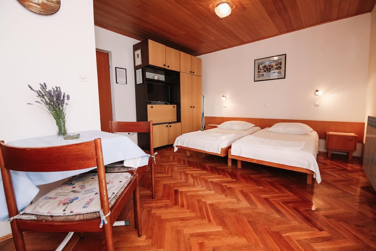 Una habitación con dos camas, una mesa de madera con un florero, una kitchenette y un piso de madera con patrón de espiga.