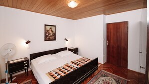 A bedroom with a wooden ceiling, a bed with a patterned blanket, a wall-mounted fan, a framed picture, and a wooden door.