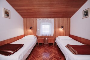 A room with two single beds, wooden panel walls, a wooden ceiling, a window with a sheer curtain, and two wall-mounted lamps.