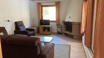 Ein Wohnzimmer mit einer Couch, einem Fernseher auf einem Holzboden, einem Kamin und einem Fenster mit Vorhängen.