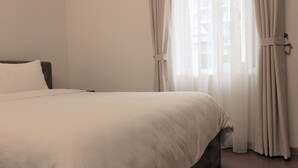 Ein ordentlich bezogenes Bett mit weißen Laken in einem Zimmer mit großem Fenster, das mit durchsichtigen Vorhängen drapiert ist.