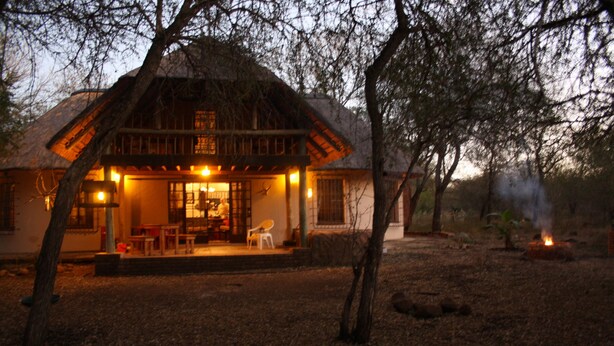 A rustic building with a thatched roof, a porch with seating, and a visible fire pit in the yard. - Sadadu (Nkomazi)