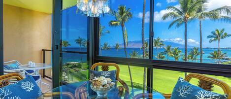 A balcony with wicker furniture, a glass table, and a view of palm trees and the ocean.
