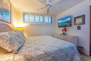 Un dormitorio con cama, ventilador de techo, televisión, ventana con persianas e imagen colgada en la pared.