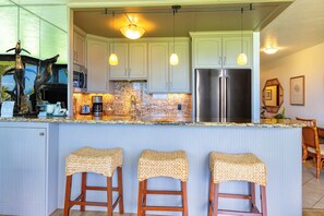 Una cocina con armarios blancos, un refrigerador de acero inoxidable y una encimera de granito. Hay tres taburetes de mimbre y una barra.