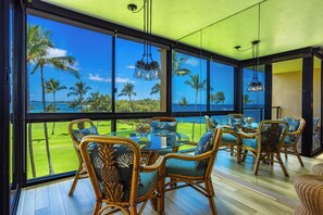 A spacious room with large windows offering a view of the ocean and palm trees. There is a dining area with wicker furniture and a glass table.