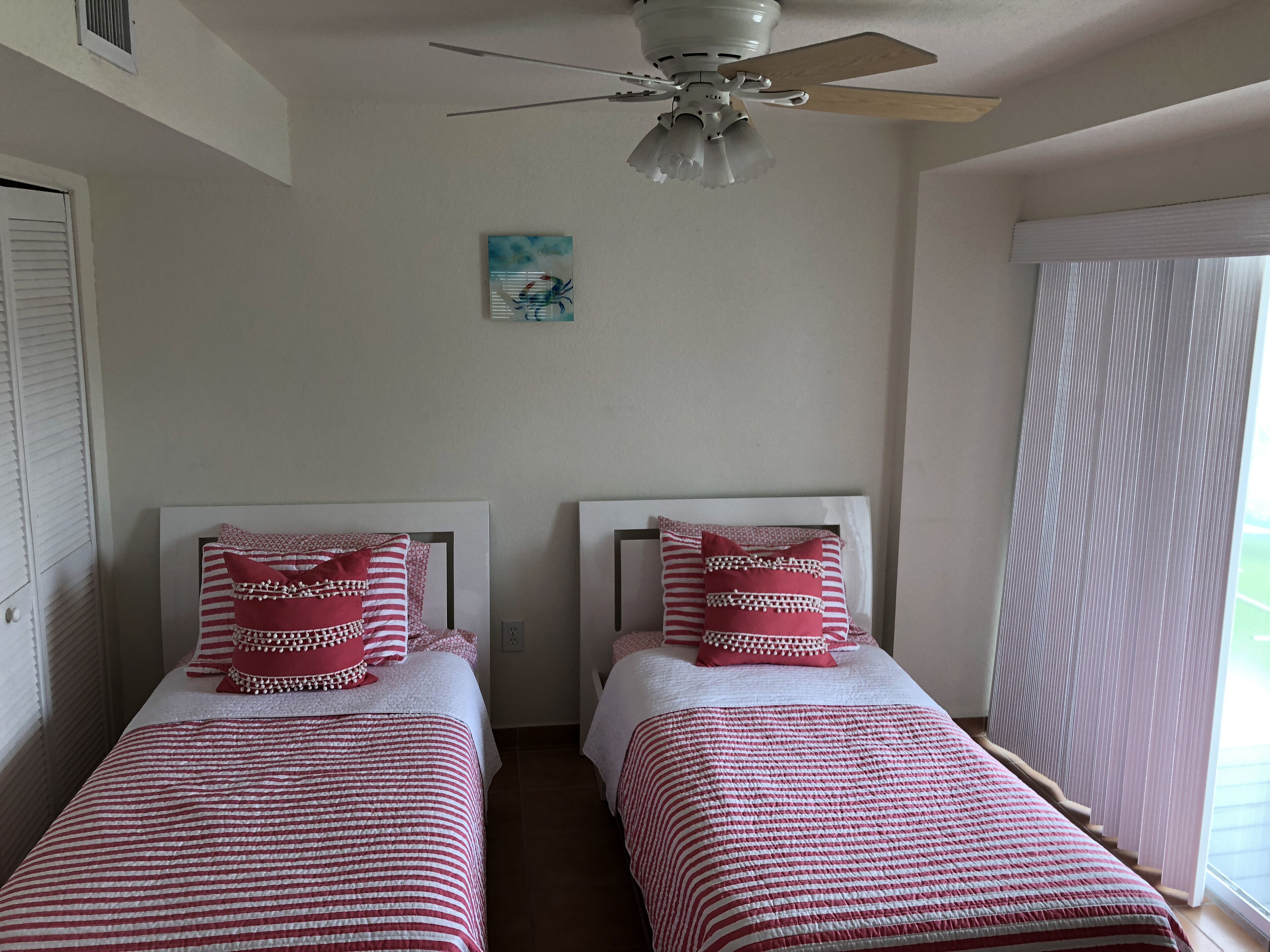 A bedroom with two beds, a ceiling fan, a window with blinds, and a small painting on the wall.