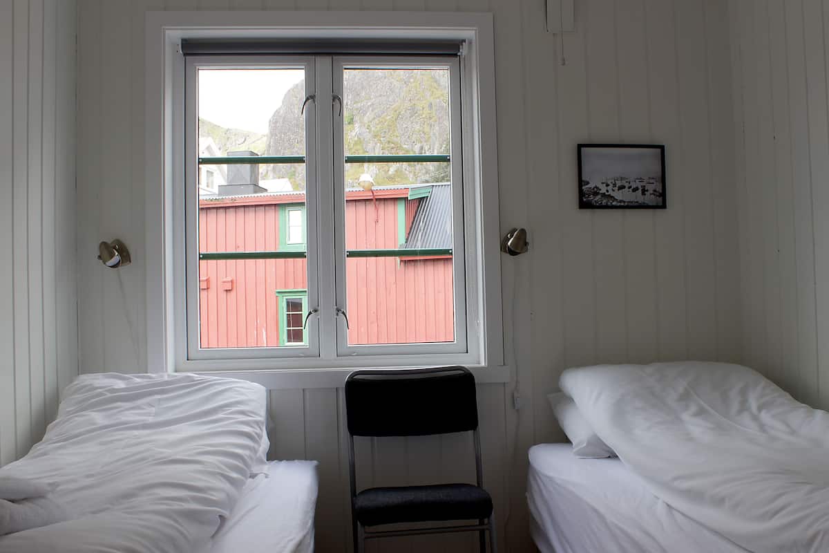 A room with two single beds, a chair, a window with a view of a red building, and a framed picture on the wall.